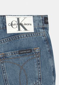 Mellanmörka blå denimjeans med en vit etikett med texten "Calvin Klein Jeans" och en svart tagg på bakfickan. Standard sömdetalj.