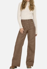 Pantalons larges marron en tissu texturé, avec une taille haute, des poches avant et des coutures contrastées. Associés à un pull léger.