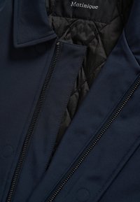 Gros plan d'une veste matelassée bleu marine avec doublure noire et fermeture éclair visible, affichant le nom de la marque "Matinique" sur le col intérieur.