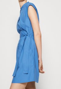 Blauwe katoenen jurk met korte mouwen, knoopsluiting aan de voorkant en een ceintuur in de taille. Gladde textuur en tot op de knie.