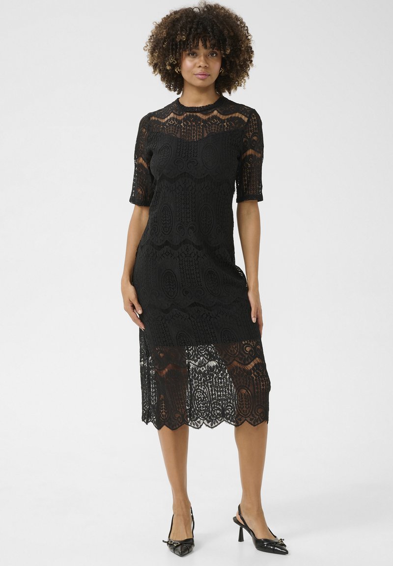 Vestido de encaje negro con mangas cortas, que presenta patrones intrincados, un corpiño ajustado y un dobladillo ondulado, combinado con zapatos de tacón negros.