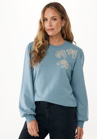 Světle modrá mikina s vypouklými rukávy a dekorativním květinovým vyšíváním ve stříbrné a béžové barvě. Vyrobená z měkké tkaniny, s kulatým výstřihem.