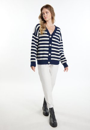 Woman wearing a navy and white striped buttoned cardigan, white pants, and black boots, standing with one leg crossed in front.