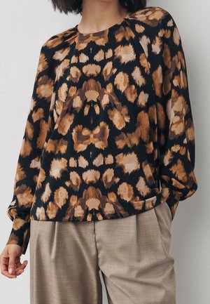Femme portant un chemisier à manches longues avec un imprimé léopard marron et noir associé à un pantalon plissé beige.