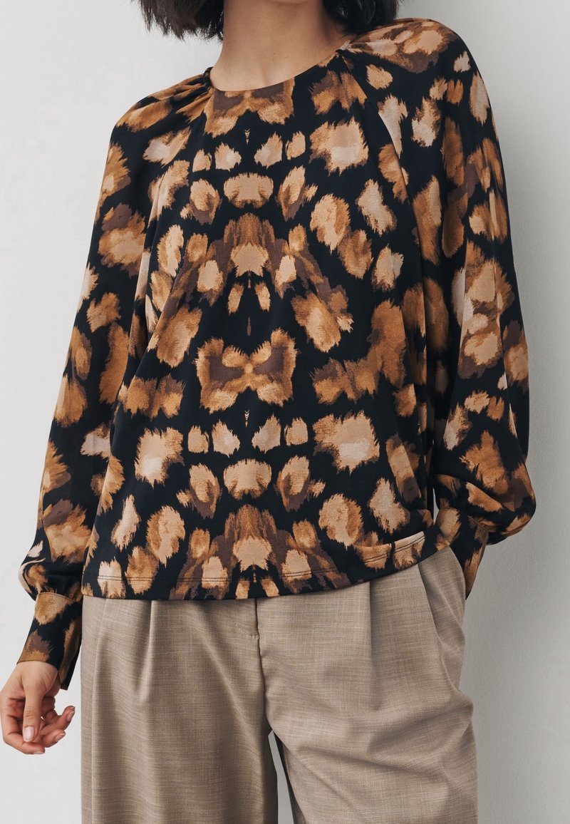 Femme portant un chemisier à manches longues avec un imprimé léopard marron et noir associé à un pantalon plissé beige.