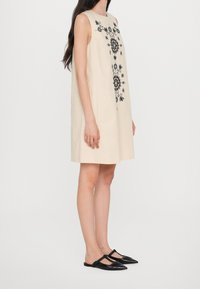 Vestido sin mangas en color beige con bordados florales negros en la parte delantera, con un corte holgado y una longitud hasta la rodilla. Combinado con bailarinas negras de punta.