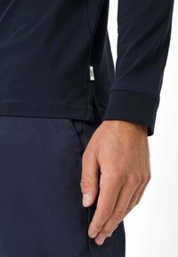 Langärmliges, marineblaues Shirt mit rundem Ausschnitt, seitlichen Schlitzen und einem Etikett am Saum. Der Stoff wirkt glatt und leicht. Nahaufnahme einer Hand, die den Rand hält.