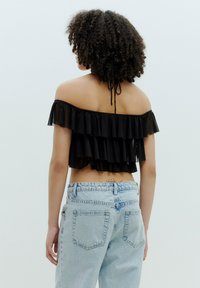 Schwarzes, schulterfreies Crop-Top mit geschichteten Rüschen und einer Schleife am Hals, kombiniert mit hellblauen, hoch taillierten Jeans.
