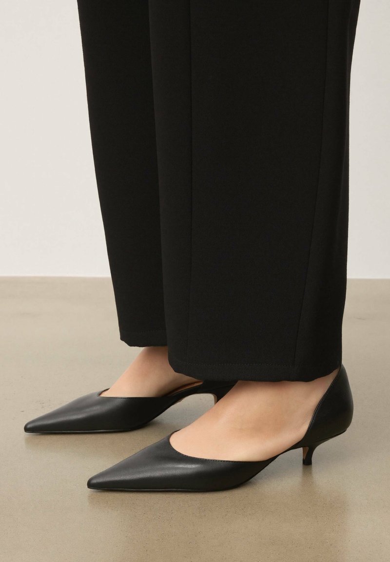 Chaussures à talons hauts en cuir noir avec bout pointu et côtés découpés. Talon aiguille fin pour ajouter de la hauteur. Portées avec un pantalon noir à jambes larges.