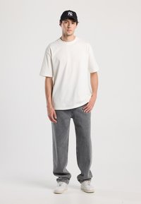 Camiseta blanca de algodón con un corte relajado, combinada con jeans de mezclilla gris. Una gorra negra con logo y zapatillas blancas completan el look.