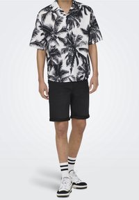 Camisa de manga corta en blanco y negro con estampado de palmeras combinada con pantalones cortos negros. Los accesorios incluyen calcetines a rayas y zapatillas en blanco y negro.