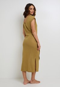 Abito midi verde oliva con schiena drappeggiata, maniche corte, spacco laterale e tessuto testurizzato. Modella in piedi a piedi nudi, con la schiena leggermente rivolta indietro.