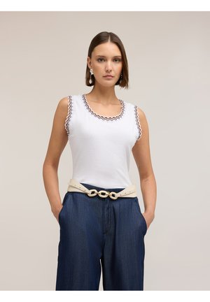 Femme portant un haut blanc sans manches avec des bords volantés, un pantalon bleu à jambes larges, une ceinture beige avec un détail chaîne et des boucles d'oreilles remarquables.