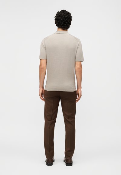 Άντρας με σγουρά σκούρα μαλλιά, ντυμένος με μπεζ κοντομάνικο πόλο, καφέ παντελόνια και σκούρα παπούτσια, στέκεται γυρισμένος προς τα πίσω σε λευκό φόντο.