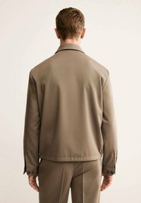 Giacca esterna marrone realizzata in tessuto liscio, con colletto classico, polsini con bottoni e design dalla vestibilità rilassata. La vista posteriore mostra uno stile minimalista.