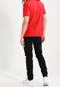 Červené polo tričko s krátkými rukávy a bočními rozparky, v kombinaci s černými úzkými džínami a bílými teniskami, pohled zezadu.