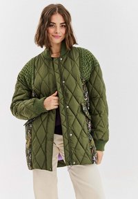 Prošívaná zelená bunda s pletenými zelenými detaily, zapínáním na druky a bočními panely s květinovým vzorem. Má žebrované manžety a volný střih.