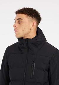 Chaqueta de plumas negra con cuello alto, cierre de cremallera y bolsillo en el pecho con cremallera; la tela parece suave con un acabado mate.