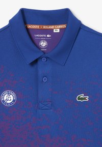 Blå pikétröja med slim fit, med ett texturerat mönster och lila accenter, märkt med Lacoste-logotypen och Roland Garros-emblemet.
