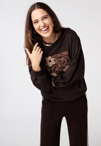 Vrouw die lacht, gekleed in een donkerbruin, gebloemd geborduurd sweatshirt, bijpassende broek met zijstrepen, gouden creolen en een gelaagde sterketting.