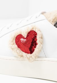 Vita sneakers med mjuk beige kant och ett stort rött paljett-hjärta på sidan. Platt sula, texturerad ovandel och snörning.