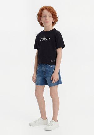 Persoană tânără cu păr roșcat în picioare, purtând un tricou negru Nike, pantaloni scurți din denim și adidași albi, pe un fundal alb simplu.