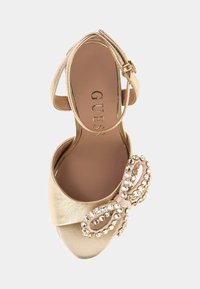 Goldene Sandale mit offener Zehenpartie, Knöchelriemen und Schnalle, mit einem mit Strass verzierten Bogen am vorderen Riemen, Guess-Logo auf der Innensohle.