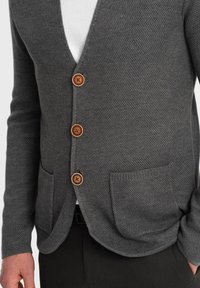 Gråtexturerad kofta med V-ringning, tre bruna knappar framtill och två framfickor. Bärs över en vit skjorta.