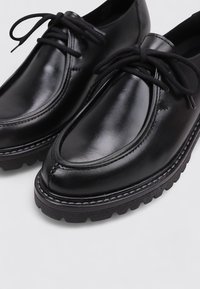 Zapatos de piel negra con un acabado liso, puntera redonda y suela de goma gruesa. Presenta un diseño cosido y cordones negros.