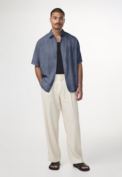 Chemise à manches courtes, de couleur bleu foncé, avec une coupe décontractée, portée sur un débardeur noir. Un pantalon large de couleur crème et des sandales noires complètent le look.