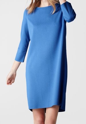 Frau trägt ein knielanges, langärmeliges blaues Kleid mit rundem Ausschnitt und steht vor einem einfarbigen hellen Hintergrund.