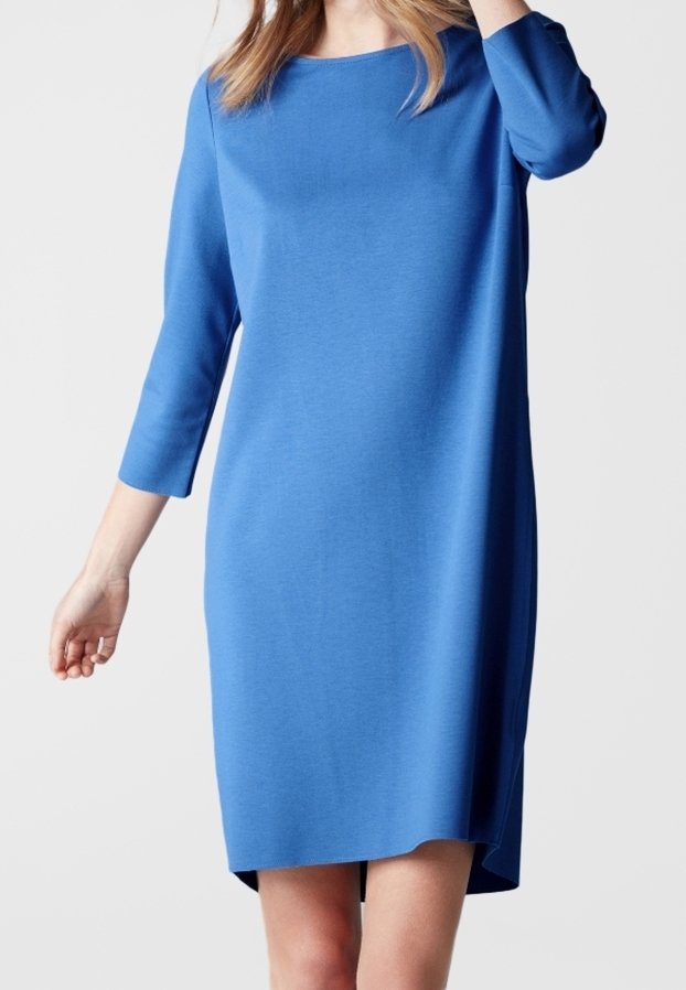 Femme portant une robe bleue mi-longue à manches longues avec un col rond, debout devant un fond clair uni.