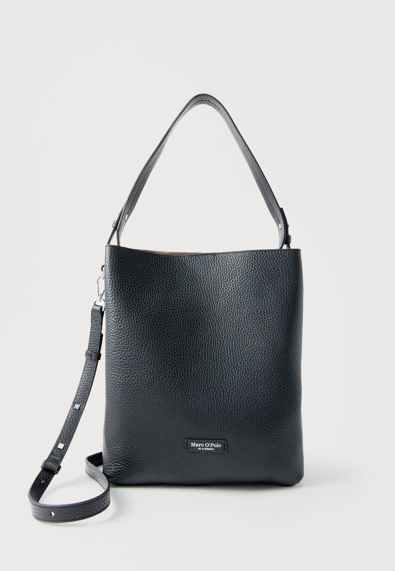 Μαύρη υφασμάτινη δερμάτινη τσάντα ώμου με ρυθμιζόμενο ιμάντα και μικρό λογότυπο στο μπροστινό μέρος, όρθια σε ουδέτερο φόντο.