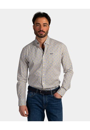 Man draagt een patroonvolle, langärmelig overhemd met knopen, ingestopt in een blauwe spijkerbroek met een zwarte riem, en staat met één hand in de zak tegen een witte achtergrond.