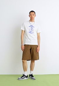 White graphic t-shirt with blue print, brown shorts, and black sneakers. Includes a simple chain necklace and sporty white socks.