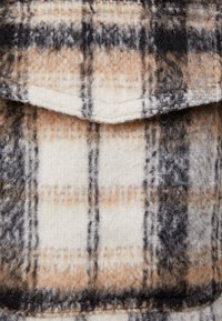 Vlněná károvaná tkanina v krémové, hnědé a černé barvě. Vyznačuje se jemnou strukturou, klopou kapsy, vodorovnými a svislými liniemi a jemnými barevnými odstíny.