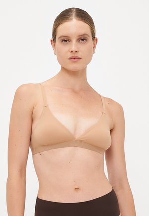 Vrouw met een lichte huid en lichtbruin haar dat naar achteren is gekamd, gekleed in een nude kleurige driehoeksbeha en donkerbruine high-waisted onderkant, tegen een witte achtergrond.