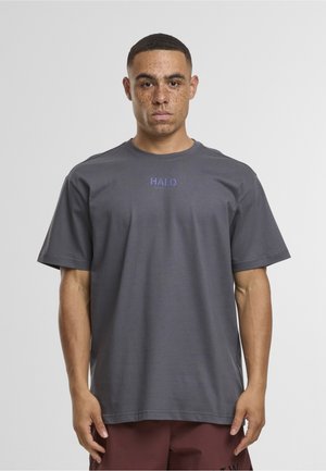 Homme aux cheveux courts et taches de rousseur portant un t-shirt gris ample marqué "HALO" et un short rouge foncé, debout devant un fond uni.