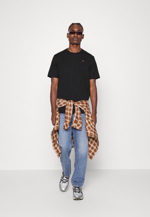 Uomo che indossa una maglietta nera, una camicia a quadri marrone legata in vita, jeans blu, sneaker argentate e occhiali da sole neri, in piedi davanti a uno sfondo bianco.