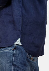 Marineblauw linnen shirt met een ronde zoom, gelaagd over een licht bloemenpatroon. Gepaard met blauwe denim jeans.