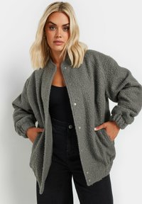 Grå fleecejacka med en mjuk, strukturerad yta; har tryckknappar, sänkta axlar och sidofickor för komfort och enkel användning.