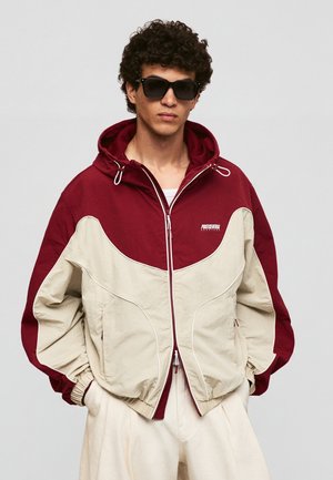 Ung mand med krøllet hår, iført mørke solbriller, en rød og beige hættejakke med lynlås og lyse bukser mod en neutral baggrund.