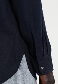Camicia di lino blu navy con maniche lunghe, spacco laterale e polsini con bottone, presentante un bottone viola. Tessuto testurizzato con una vestibilità comoda.