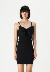 Robe noire moulante ornée d'un grand nœud au niveau de la poitrine, avec des bretelles fines et une texture lisse. Le design est minimaliste et ajusté.