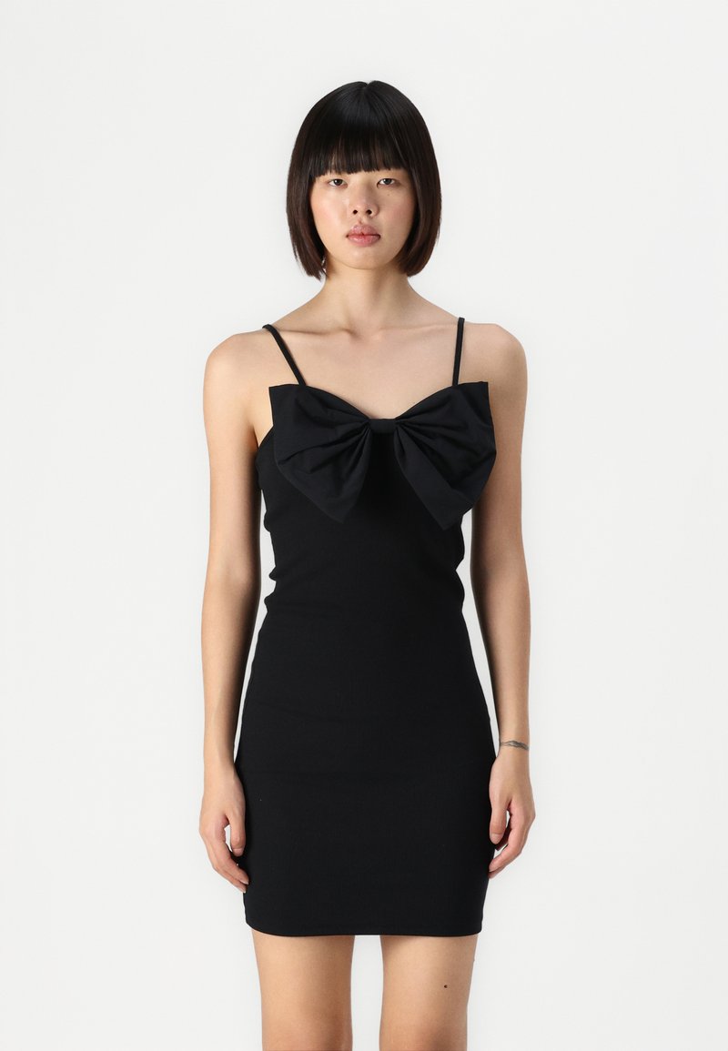 Robe noire moulante ornée d'un grand nœud au niveau de la poitrine, avec des bretelles fines et une texture lisse. Le design est minimaliste et ajusté.