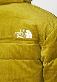 Olivgrön corduroy-tygpufferjacka med quiltad design och vitt broderat logo på vänster axel. Mjuk textur och randsydd mönster.