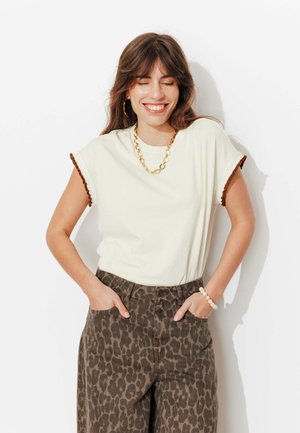 Femme souriante aux cheveux bruns ondulés porte un haut crème avec une bordure marron, un collier chaîne en or, un pantalon imprimé léopard, des boucles d'oreilles anneaux et un bracelet de perles.