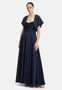 Granatowa satynowa sukienka z opadającym dekoltem, krótkimi falbaniastymi rękawami i pełną spódnicą, która opada do podłogi, w połączeniu z minimalistycznym obuwiem.