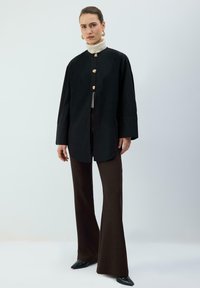 Giacca nera oversize con bottoni dorati, maniche lunghe e texture liscia, abbinata a pantaloni marroni svasati e scarpe nere appuntite.
