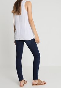 Mujer vistiendo una blusa sin mangas blanca, jeans ajustados azul oscuro y sandalias planas beige, de pie contra un fondo claro y sencillo.